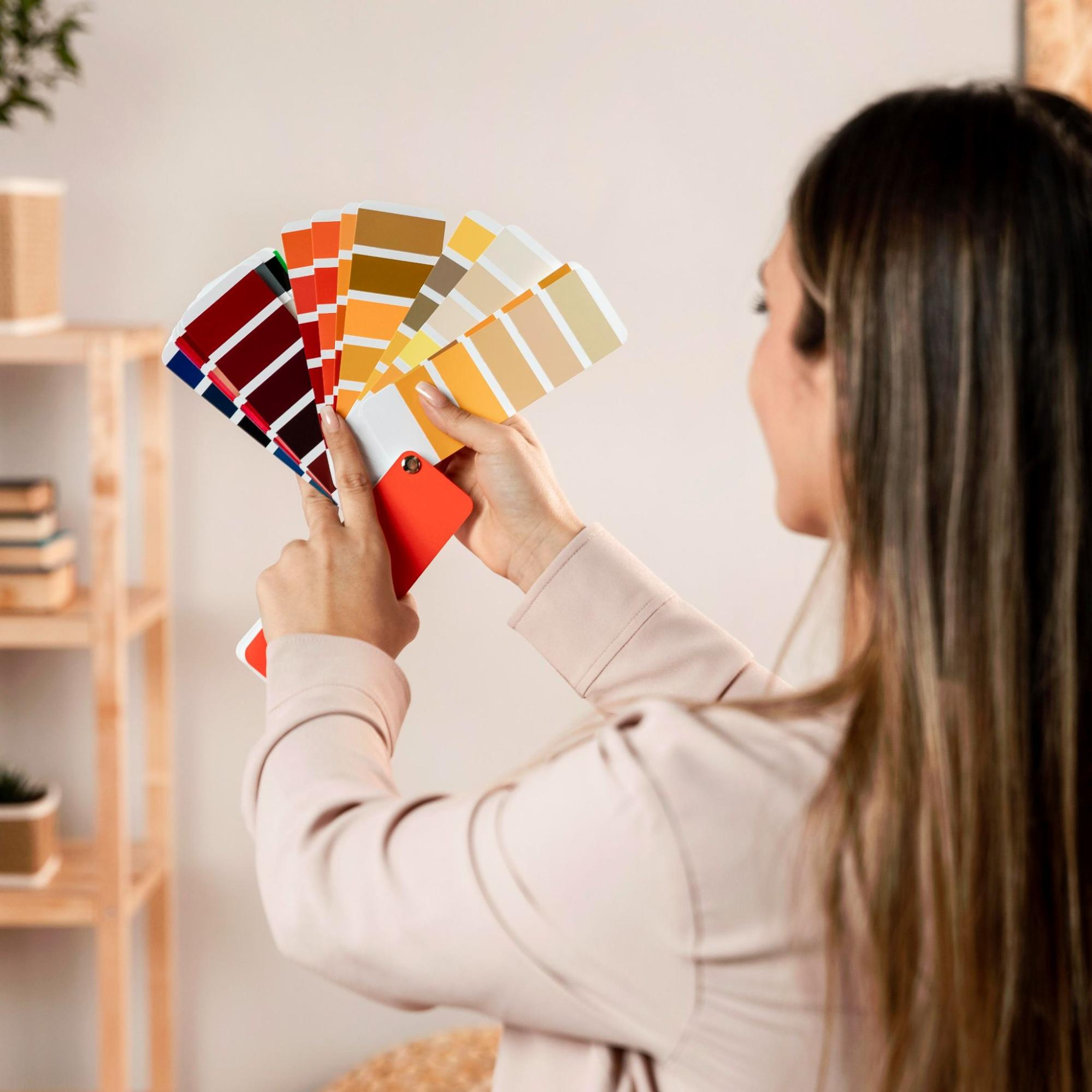 Foto em ângulo lateral de uma mulher de cabelos castanhos longos e casaco bege segurando um leque aberto com amostras de cores de tinta. Ao fundo, desfocado, vê-se um ambiente interno iluminado com uma estante de madeira clara contendo livros e vasos.
Foto em ângulo lateral de uma mulher de cabelos castanhos longos e casaco bege segurando um leque aberto com amostras de cores de tinta. Ao fundo, desfocado, vê-se um ambiente interno iluminado com uma estante de madeira clara contendo livros e vasos.