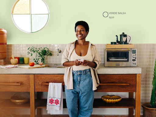 Uma mulher negra sorridente com cabelos curtos, vestindo uma camisa bege aberta sobre um top de tricô e calça jeans, está de pé em uma cozinha rústica de conceito aberto, segurando um smartphone. Ela está apoiada em um balcão de concreto com base de madeira. A parede atrás dela é pintada em um tom suave de verde-claro. O balcão tem uma pia, um pequeno forno elétrico, uma cafeteira e vários itens de cozinha, como um filtro de barro, tábua de corte com vegetais, vasos e um cacto grande à direita. Uma mulher negra sorridente com cabelos curtos, vestindo uma camisa bege aberta sobre um top de tricô e calça jeans, está de pé em uma cozinha rústica de conceito aberto, segurando um smartphone. Ela está apoiada em um balcão de concreto com base de madeira. A parede atrás dela é pintada em um tom suave de verde-claro. O balcão tem uma pia, um pequeno forno elétrico, uma cafeteira e vários itens de cozinha, como um filtro de barro, tábua de corte com vegetais, vasos e um cacto grande à direita.