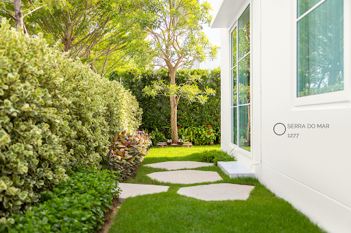 Área externa de uma casa moderna com parede pintada em tom off-white suave. À esquerda, há um jardim bem cuidado com gramado verde, caminho de pisantes de pedra clara e arbustos densos, além de grandes janelas de vidro refletindo a vegetação ao redor.
Área externa de uma casa moderna com parede pintada em tom off-white suave. À esquerda, há um jardim bem cuidado com gramado verde, caminho de pisantes de pedra clara e arbustos densos, além de grandes janelas de vidro refletindo a vegetação ao redor.