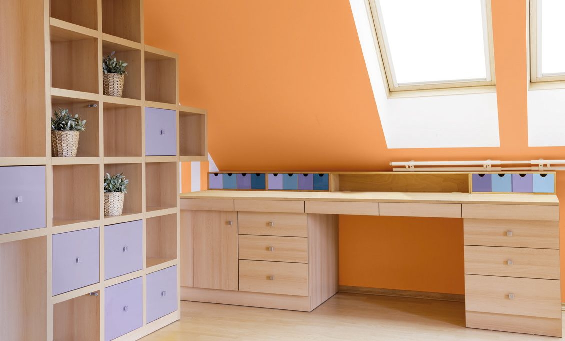 Ambiente interno com estantes e mesa de madeira clara em um quarto com parede laranja. As estantes possuem nichos quadrados, alguns com plantas e outros com gavetas lilás. Ambiente interno com estantes e mesa de madeira clara em um quarto com parede laranja. As estantes possuem nichos quadrados, alguns com plantas e outros com gavetas lilás.