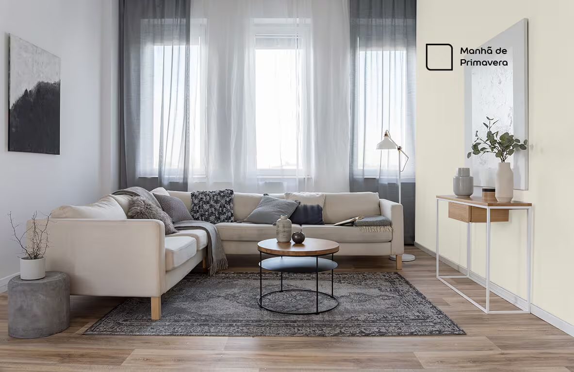 Uma sala de estar decorada com paredes pintadas em um tom de bege claro, chamado 'Manhã de Primavera'. O ambiente tem um sofá seccional claro com almofadas cinzas e estampadas, uma mesa de centro de madeira, um tapete cinza com estampa geométrica no chão, uma grande janela com persianas, um espelho de corpo inteiro e uma mesa de console estreita com vasos decorativos.