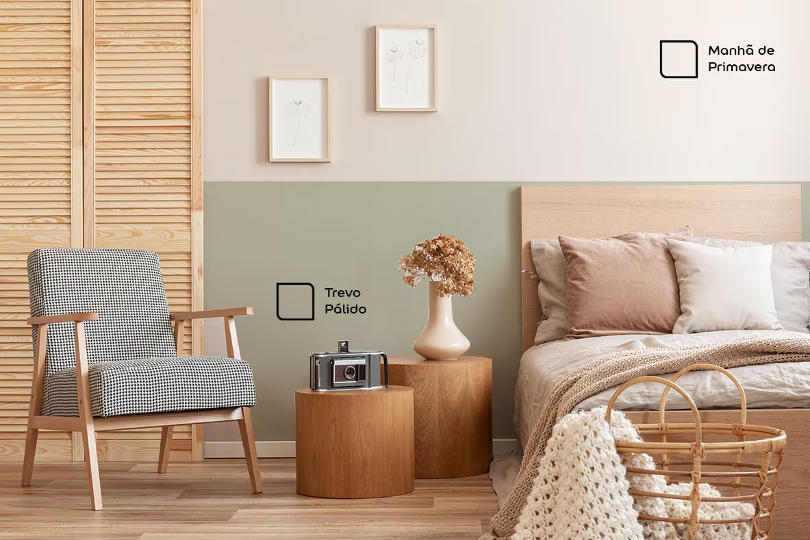Um quarto moderno e aconchegante com uma parede de dois tons, pintada com as cores 'Manhã de Primavera' (parte superior) e 'Trevo Pálido' (parte inferior). O ambiente tem uma cama com cabeceira de madeira e roupa de cama neutra, uma poltrona de tecido xadrez, mesinhas de cabeceira redondas de madeira clara e quadros decorativos na parede.