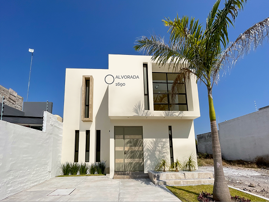 Fachada de uma casa moderna de dois andares com paredes na cor creme ou bege claro. A casa possui janelas retangulares escuras e uma grande porta de entrada com detalhes em cor bege e marrom. Há plantas baixas na frente da casa, uma palmeira à direita e o chão é de cimento. Há um número de endereço e um nome na fachada.