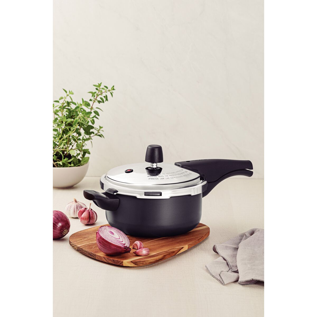 Uma panela de pressão Tramontina Vancouver preta sobre uma tábua de madeira, com uma cebola roxa e alho descascado ao lado. A panela de pressão tem uma alça longa e uma alça menor, com uma válvula de segurança na tampa prateada. Uma panela de pressão Tramontina Vancouver preta sobre uma tábua de madeira, com uma cebola roxa e alho descascado ao lado. A panela de pressão tem uma alça longa e uma alça menor, com uma válvula de segurança na tampa prateada.