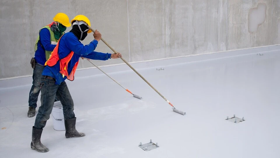 Dois trabalhadores da construção civil usando capacetes amarelos, coletes refletivos e botas aplicam manta líquida branca impermeabilizante no piso de uma laje com rolos de pintura de cabo longo. A superfície do chão está coberta pelo produto branco e uniforme ao lado de uma parede de concreto cinza. Dois trabalhadores da construção civil usando capacetes amarelos, coletes refletivos e botas aplicam manta líquida branca impermeabilizante no piso de uma laje com rolos de pintura de cabo longo. A superfície do chão está coberta pelo produto branco e uniforme ao lado de uma parede de concreto cinza.