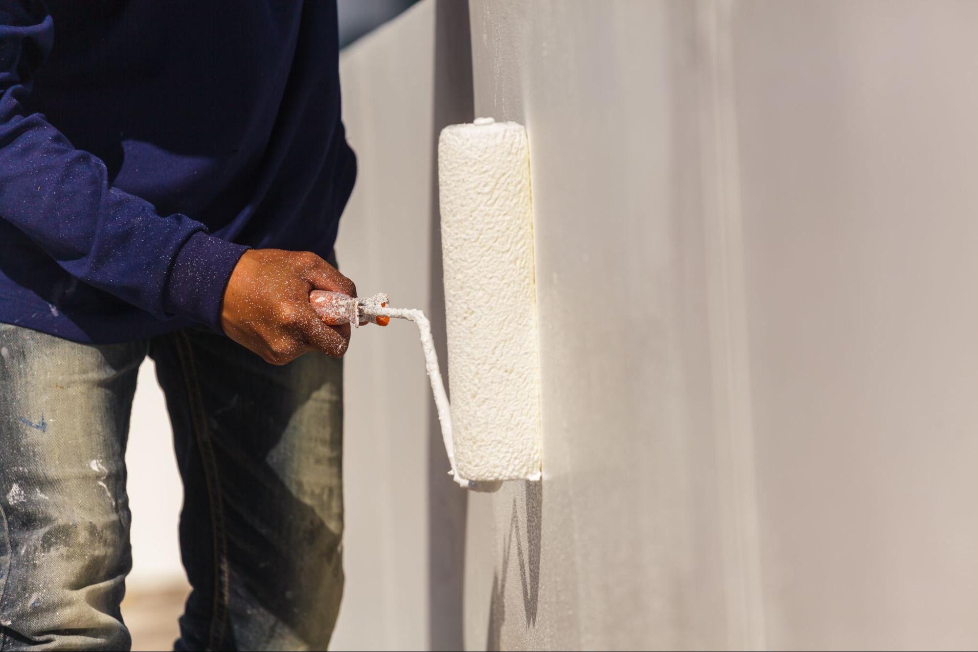 Uma pessoa vestindo camisa azul e jeans manchados de tinta, com as mãos sujas de tinta branca, está pintando uma parede ou superfície vertical de branco usando um rolo de pintura. O rolo está carregado de tinta e cobrindo a superfície, com respingos de tinta visíveis. Uma pessoa vestindo camisa azul e jeans manchados de tinta, com as mãos sujas de tinta branca, está pintando uma parede ou superfície vertical de branco usando um rolo de pintura. O rolo está carregado de tinta e cobrindo a superfície, com respingos de tinta visíveis.