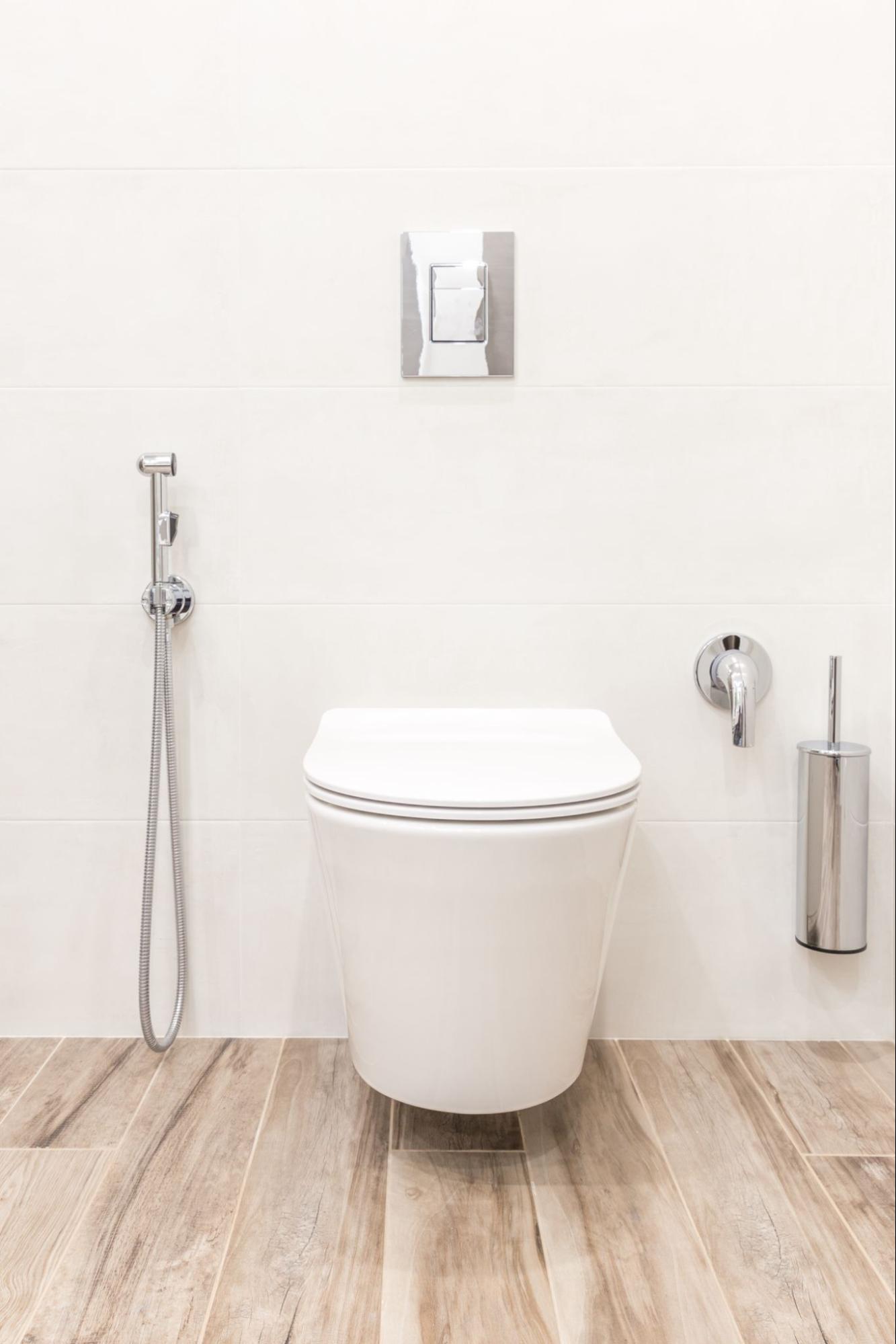 Vaso sanitário suspenso branco, instalado em uma parede de azulejos brancos, com piso de madeira clara. Acima do vaso, há uma descarga embutida cromada. À esquerda, uma ducha higiênica cromada e à direita, um porta-escova de vaso e registro de água, ambos cromados.