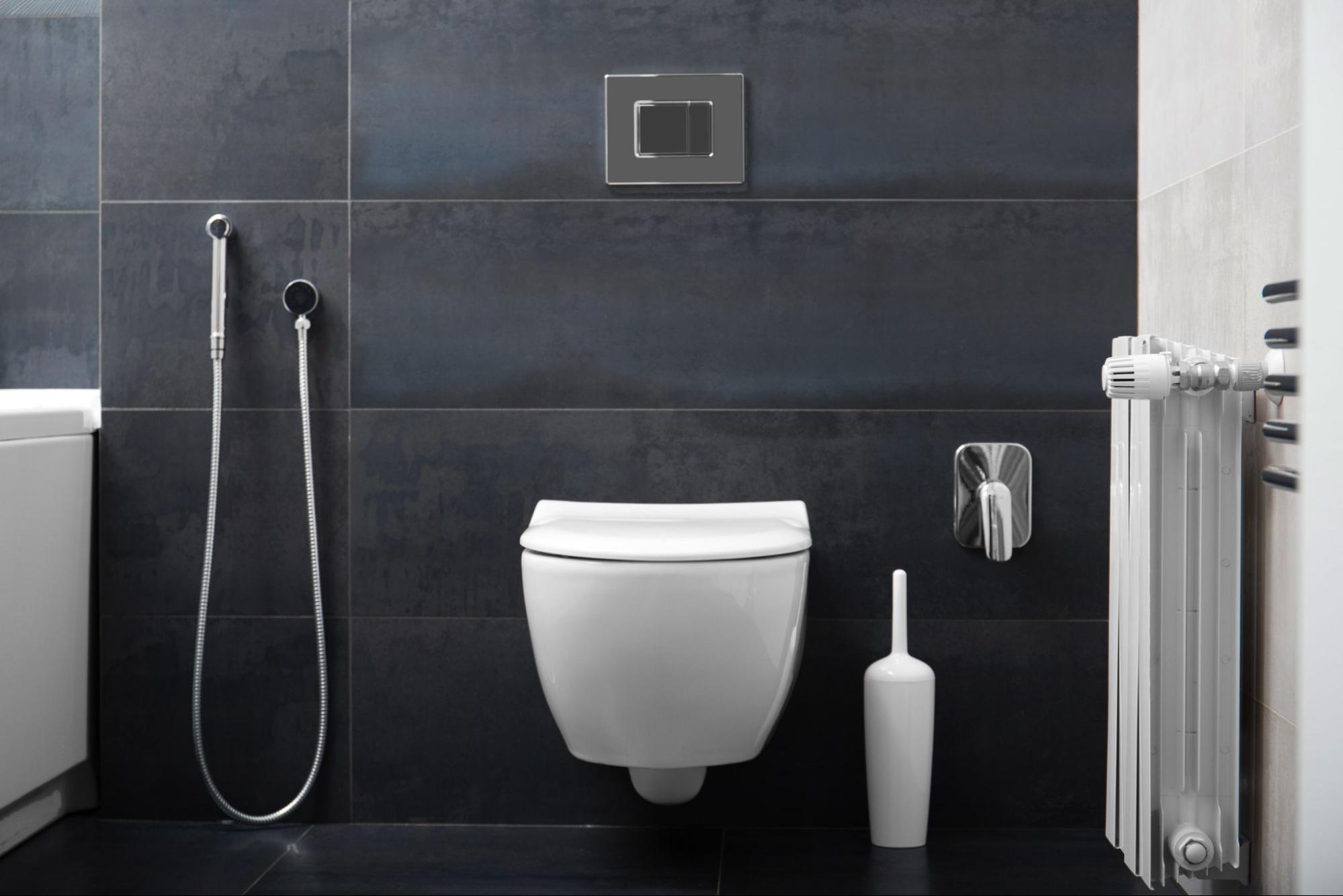 Vaso sanitário branco suspenso em um banheiro com revestimento de parede em azulejos cinza escuro. À esquerda, uma ducha higiênica cromada presa à parede. Acima do vaso, uma placa de descarga retangular cromada. À direita, um porta-escova de vaso sanitário branco e um toalheiro aquecido branco na parede.
