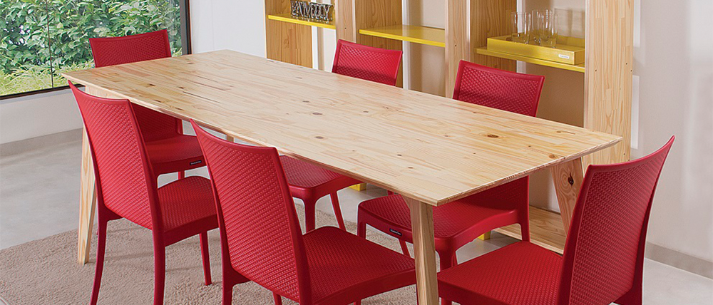 Sala de jantar moderna com mesa de madeira clara e seis cadeiras Tramontina vermelhas texturizadas. Ao fundo, há uma estante de madeira com prateleiras amarelas e uma grande janela vista para um jardim verde. No canto inferior esquerdo, uma tigela branca com texto em português sobre o produto.