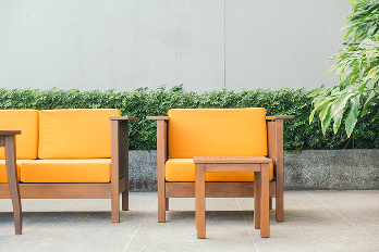Conjunto de móveis para área externa em madeira clara com estofados amarelos, composto por um sofá de dois lugares, uma poltrona e uma mesa de centro quadrada, sobre um piso de pedra clara em frente a uma parede cinza com vegetação.