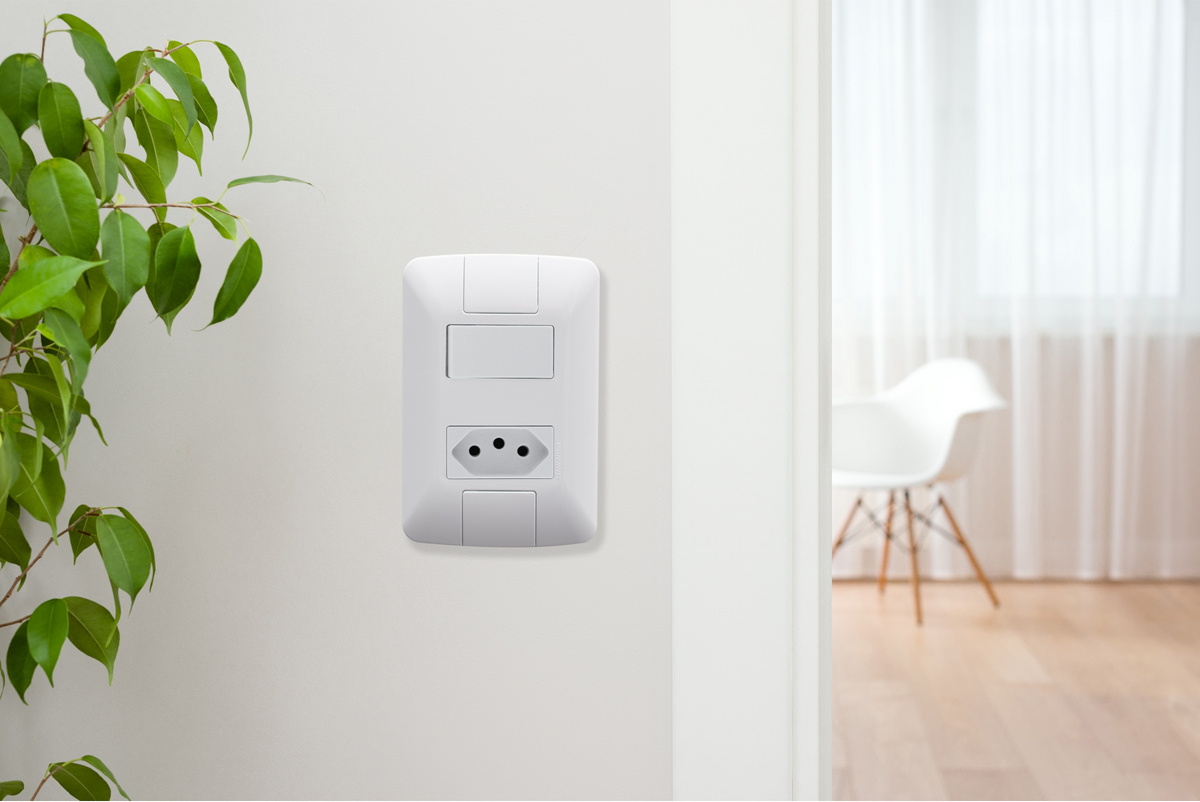 Uma placa de parede branca com um interruptor simples na parte superior e uma tomada de três pinos na parte inferior, instalada em uma parede clara. À esquerda, uma planta verde, e à direita, um vislumbre de um ambiente com uma cadeira branca e cortinas claras