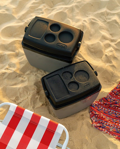 Dois coolers ou caixas térmicas da marca MOR, com corpo cinza e tampa preta, em uma superfície de areia. A tampa de cada cooler possui porta-copos. No canto inferior, parte de uma cadeira de praia de alumínio com tecido listrado em vermelho e branco é visível.