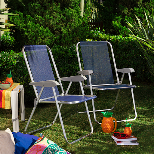 Duas cadeiras de praia dobrável, com estrutura de alumínio prateado e assento de tecido na cor azul-marinho. As cadeiras estão em um gramado verde, cercado por arbustos. Ao lado, uma pequena mesa branca com morangos e, no chão, itens decorativos de abacaxi e óculos escuros.