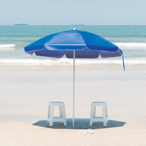 Um guarda-sol grande na cor azul com acabamento branco, fincado na areia branca da praia. Duas banquetas ou banquinhos de plástico brancos estão colocados sob o guarda-sol. Ao fundo, o mar azul com ondas suaves e navios no horizonte.