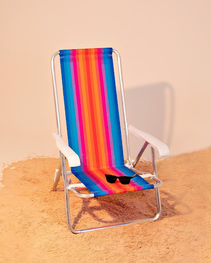 Uma cadeira de praia reclinável, com estrutura em aço prateado e assento de tecido listrado em tons vibrantes de laranja, rosa, azul e amarelo. A cadeira está posicionada sobre uma superfície de areia, e um óculos de sol preto está sobre o assento.