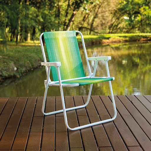 Uma cadeira de praia dobrável com estrutura de aço pintado de branco e assento de tecido listrado em tons de verde e amarelo. A cadeira está em uma posição semi-inclinada, em um deque de madeira sobre um lago. Ao fundo, árvores e vegetação densa.