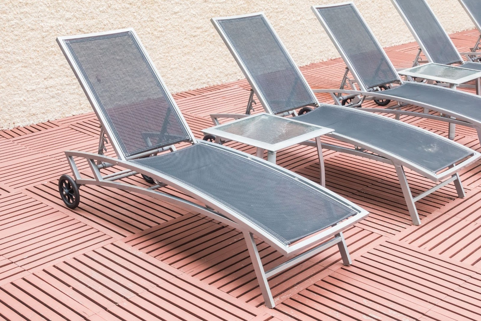 Um conjunto de espreguiçadeiras com estrutura de alumínio prateado e assento de tecido cinza, dispostas em um deque de madeira marrom-avermelhada. Entre algumas espreguiçadeiras, há mesas pequenas com tampo de vidro. Ao fundo, a areia clara é visível.