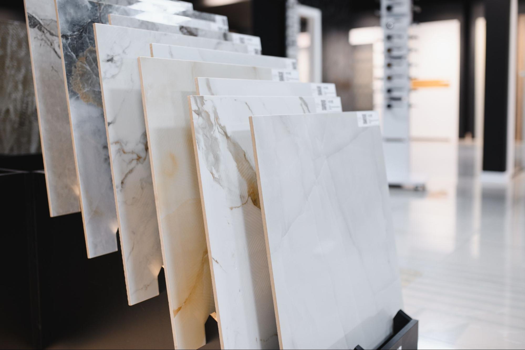 Mostruário de diversas placas de porcelanato de grandes formatos, inclinadas em um suporte preto em um showroom. As placas exibem diferentes texturas e padrões, incluindo simulações de mármore (calacata) com veios cinza e dourados, e tons de branco e cinza. O ambiente é iluminado e moderno.