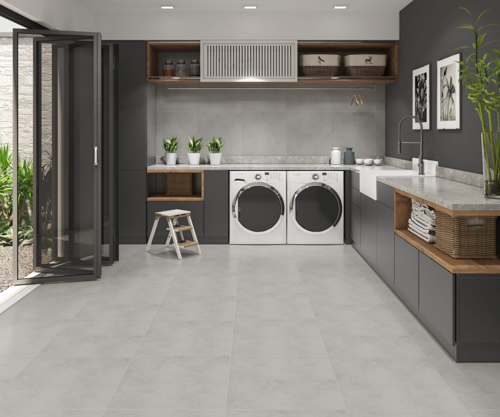 Área de serviço moderna com piso revestido por porcelanato cimento queimado em grandes formatos retificados. O ambiente possui paredes cinzas, armários escuros com detalhes em madeira clara, duas máquinas de lavar frontais brancas, uma pia branca, uma pequena escada de madeira branca e uma porta de vidro dobrável aberta para uma área externa com plantas.