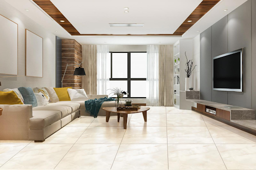 Sala de estar moderna e aconchegante revestida com porcelanato marmorizado em tom bege-claro e acabamento brilhante no piso. À esquerda, um sofá em L bege com almofadas amarelas e azuis, e uma mesa de centro de madeira. À direita, um painel cinza com televisão e rack suspenso. O teto possui sancas de madeira com iluminação embutida e grandes janelas trazem luz natural ao fundo. Sala de estar moderna e aconchegante revestida com porcelanato marmorizado em tom bege-claro e acabamento brilhante no piso. À esquerda, um sofá em L bege com almofadas amarelas e azuis, e uma mesa de centro de madeira. À direita, um painel cinza com televisão e rack suspenso. O teto possui sancas de madeira com iluminação embutida e grandes janelas trazem luz natural ao fundo.