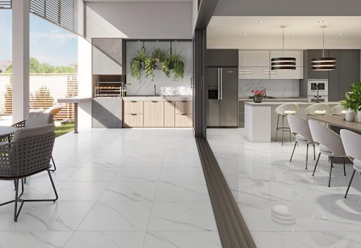 Ambiente integrado de conceito aberto, mostrando uma varanda gourmet externa e uma cozinha moderna interna, ambas revestidas com grandes placas de porcelanato marmorizado polido, imitando um piso de mármore branco com veios cinzas. As portas de correr de vidro estão totalmente abertas, conectando os espaços. A área gourmet tem churrasqueira, armários de madeira e plantas, enquanto a cozinha interna tem uma ilha central com bancos e área de jantar adjacente. Ambiente integrado de conceito aberto, mostrando uma varanda gourmet externa e uma cozinha moderna interna, ambas revestidas com grandes placas de porcelanato marmorizado polido, imitando um piso de mármore branco com veios cinzas. As portas de correr de vidro estão totalmente abertas, conectando os espaços. A área gourmet tem churrasqueira, armários de madeira e plantas, enquanto a cozinha interna tem uma ilha central com bancos e área de jantar adjacente.