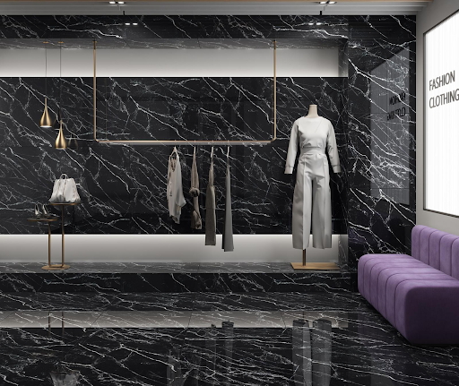 Interior de uma loja de roupas luxuosa e moderna com revestimento em porcelanato marmorizado preto e veios brancos marcantes cobrindo o piso e as paredes. À direita, há um manequim com conjunto branco e um sofá aveludado na cor roxa. Ao centro, uma arara dourada suspensa exibe algumas peças de roupa, complementada por luminárias pendentes douradas e fitas de LED embutidas. Interior de uma loja de roupas luxuosa e moderna com revestimento em porcelanato marmorizado preto e veios brancos marcantes cobrindo o piso e as paredes. À direita, há um manequim com conjunto branco e um sofá aveludado na cor roxa. Ao centro, uma arara dourada suspensa exibe algumas peças de roupa, complementada por luminárias pendentes douradas e fitas de LED embutidas.