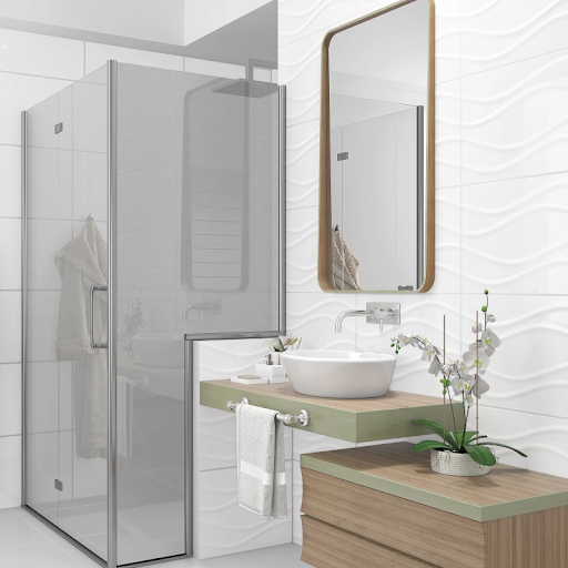 um banheiro moderno e claro. A parede de destaque é revestida com cerâmica branca em alto-relevo que simula ondas. Há um box de vidro com ferragens prateadas e um chuveiro de teto. A bancada é suspensa, com cuba de apoio branca e estrutura de madeira com detalhes em verde-claro. Um espelho retangular com moldura de madeira está sobre a bancada, e uma orquídea branca decora o espaço. 