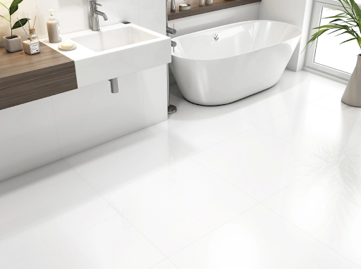 um banheiro moderno com piso e paredes revestidos com peças grandes e brancas, com acabamento brilhante. O piso reflete o ambiente. Há uma cuba de apoio branca sobre uma bancada de madeira escura suspensa, uma banheira freestanding branca no fundo e um vaso de planta à direita, próximo à janela.