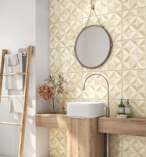 um banheiro moderno com uma parede de destaque revestida com cerâmica geométrica em tons de bege e branco, com um padrão em relevo que simula pétalas ou curvas. Há uma bancada de madeira, uma cuba branca e uma torneira de metal curvada. Um espelho redondo pendurado por uma corda, e uma escada de madeira decora a lateral, segurando toalhas.