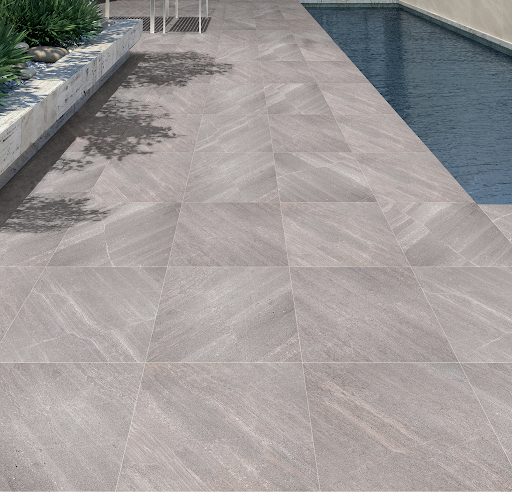 Área externa de deck com piscina coberta por piso cerâmico cinza em tom médio, com textura suave de pedra natural disposta em padrão diagonal. À esquerda, há um canteiro elevado de pedra clara com folhagens verdes e sombras de árvores projetadas no chão. À direita, uma piscina estreita de água escura segue ao longo da extensão do ambiente. Área externa de deck com piscina coberta por piso cerâmico cinza em tom médio, com textura suave de pedra natural disposta em padrão diagonal. À esquerda, há um canteiro elevado de pedra clara com folhagens verdes e sombras de árvores projetadas no chão. À direita, uma piscina estreita de água escura segue ao longo da extensão do ambiente.