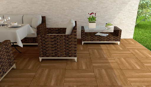 área externa com piso amadeirado em formato quadriculado e parede ao fundo revestida com cor clara. O ambiente conta com móveis de fibra trançada escura e estofado branco, incluindo poltronas, mesa de jantar posta e uma mesa de centro de vidro decorada com vasos de flores. À direita, há uma abertura com vista para um gramado verde. área externa com piso amadeirado em formato quadriculado e parede ao fundo revestida com cor clara. O ambiente conta com móveis de fibra trançada escura e estofado branco, incluindo poltronas, mesa de jantar posta e uma mesa de centro de vidro decorada com vasos de flores. À direita, há uma abertura com vista para um gramado verde.