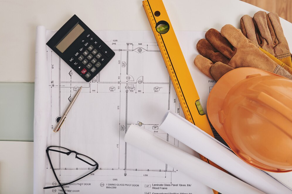 Planta baixa de projeto elétrico sobre mesa, com calculadora, caneta, óculos, luvas e capacete de segurança. Planta baixa de projeto elétrico sobre mesa, com calculadora, caneta, óculos, luvas e capacete de segurança.